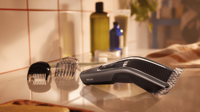 Product close-up image of a black and grey Philips hair clipper with comb attachment laying on a bathroom counter.