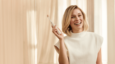 A young blonde woman wearing a white outfit holds a Philips electric toothbrush up and smiles to show whitened teeth.