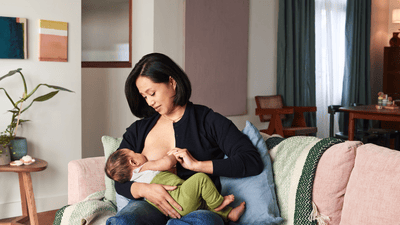 Een vrouw zit op een roze bank terwijl ze haar baby borstvoeding geeft in een felgekleurde woonkamer.