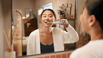 A woman with dark hair, wearing a white robe, is watching herself brush her teeth in the mirror.