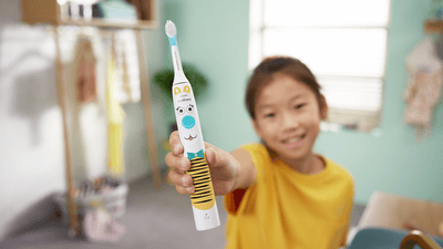 A young child wearing yellow holds their Philips kids electric toothbrush up proudly to the camera.