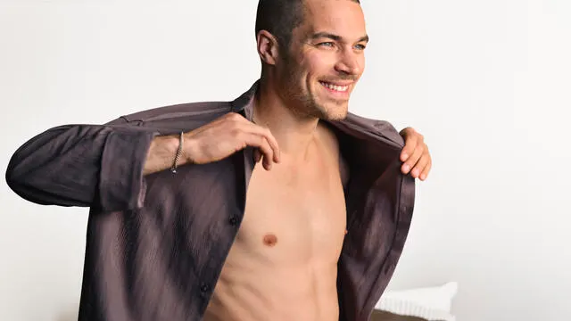 A young man wearing an unbuttoned brown shirt smiles widely.