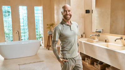 A bald man with a nicely trimmed beard stands in a stylish bathroom, wearing light-coloured clothes, and smiles at the camera.