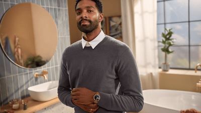A dressed man with a long stubble poses directly at the camera in a bathroom.