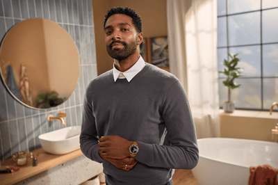 A dressed man with a long stubble poses directly at the camera in a bathroom.