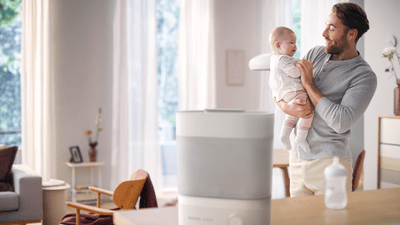 A father smiles while holding his baby in a bright living room with a Philips 2-in-1 bottle warmer and steriliser on the counter.
