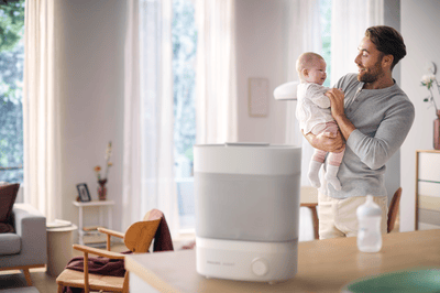 A father smiles while holding his baby in a bright living room with a Philips 2-in-1 bottle warmer and steriliser on the counter.