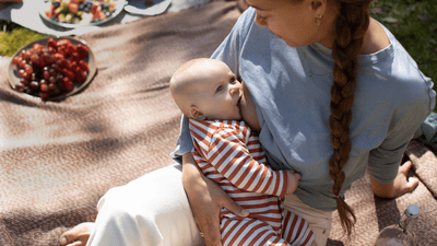 A woman breastfeeds her baby on a light brown picnic blanket beside plates of brightly coloured food.