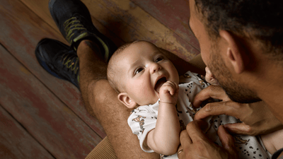 A smiling baby is lying on a man's legs, looking up joyfully as he plays with the baby.