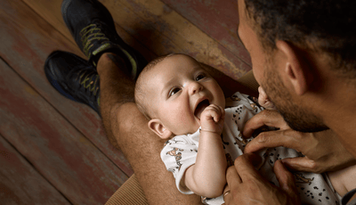 A smiling baby is lying on a man's legs, looking up joyfully as he plays with the baby.