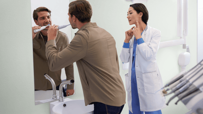 A young dental professional stands beside a young man wearing a brown shirt and brushing his teeth in the mirror.