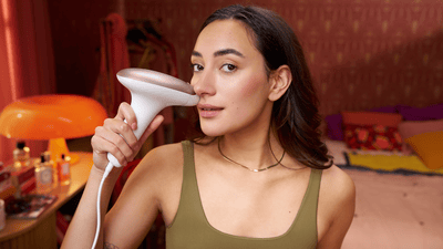 A woman uses a Philips Lumea IPL device on her face and chin hair in a bright, colourful bedroom setting.