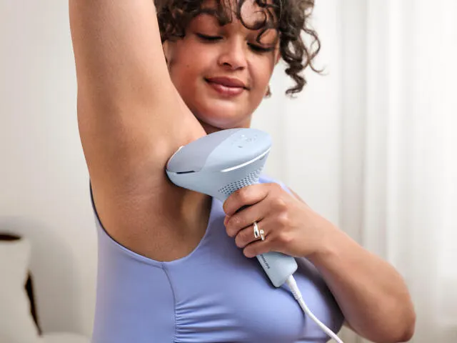 A curly-haired woman uses a Philips Lumea IPL device on her underarms.