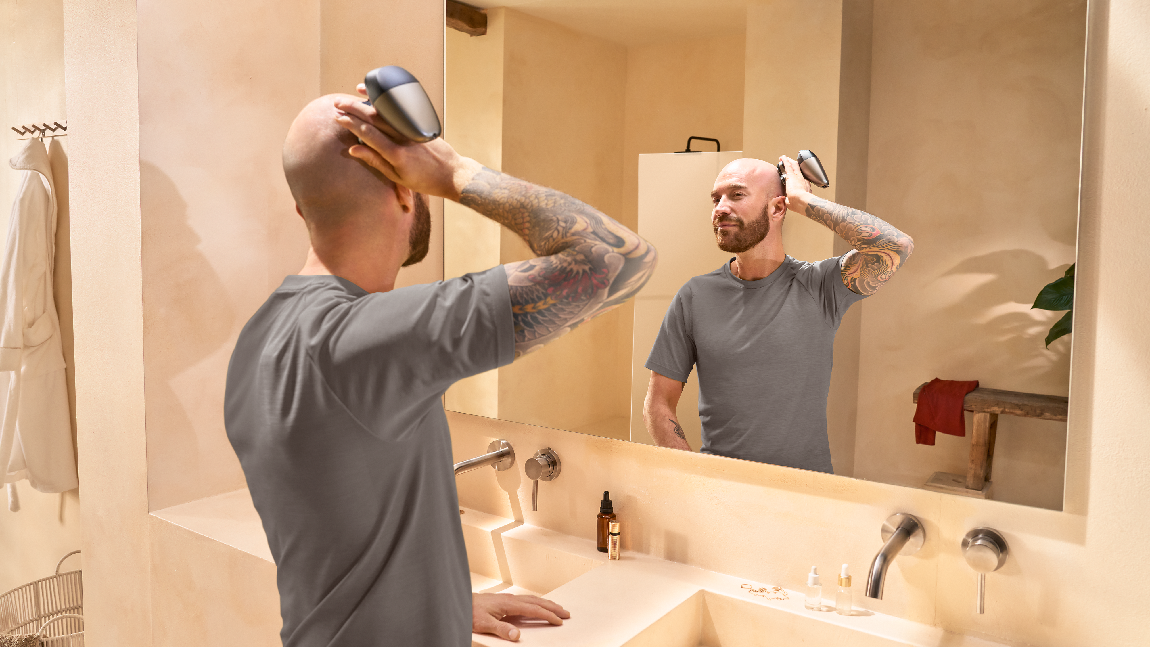 A bald man with a sleeve tattoo shaves his head with a Philips head shaver in front of a bathroom mirror.