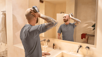A bald man with a sleeve tattoo shaves his head with a Philips head shaver in front of a bathroom mirror.