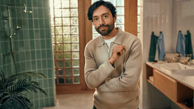 A man wearing a beige jumper with a long stubble poses directly at the camera in a bathroom.
