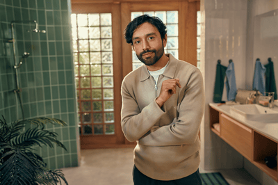 A man wearing a beige jumper with a long stubble poses directly at the camera in a bathroom.