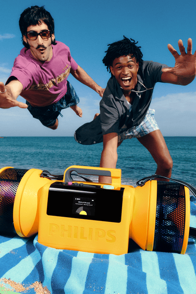 Two men jumping mid-air on a sunny beach above a yellow portable speaker on a towel,