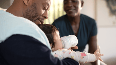 A man feeds his young infant with a Philips Avent baby bottle, while another man sits and observes.
