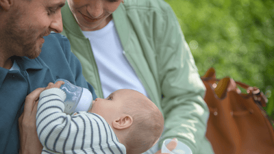 A man feeds his young infant with a Philips Avent baby bottle, while a woman watches in an outdoor setting.