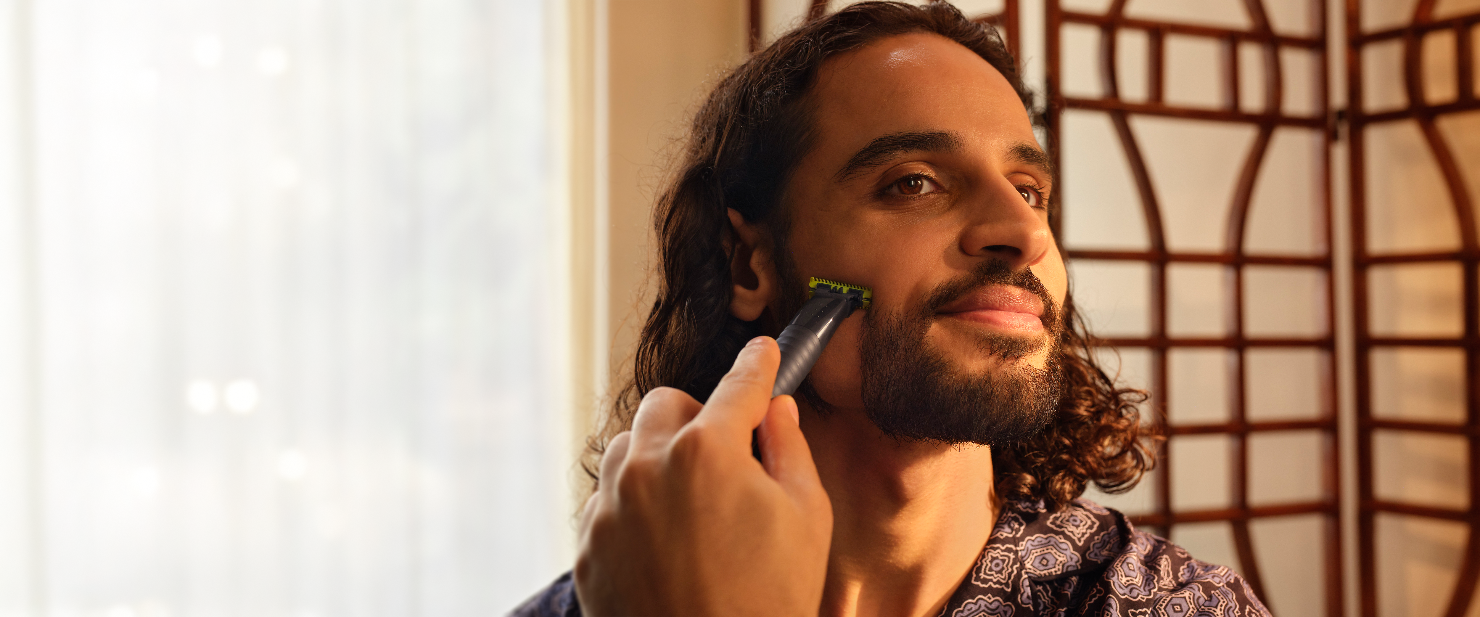 A man with curly hair smiles as he shaves his cheek with the Philips OneBlade.