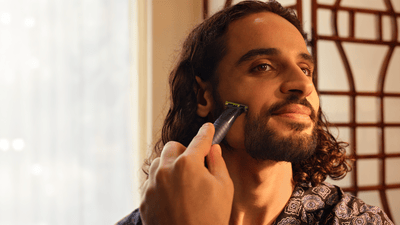 A man with curly hair smiles as he shaves his cheek with the Philips OneBlade.