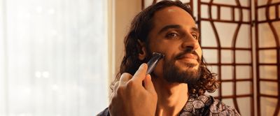 A man with curly hair smiles as he shaves his cheek with the Philips OneBlade.