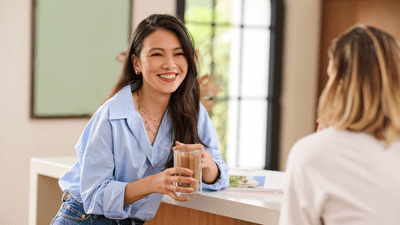A woman with dark hair, wearing a blue blouse, holds a glass of iced coffee and sits across from a blonde woman in a white blouse.