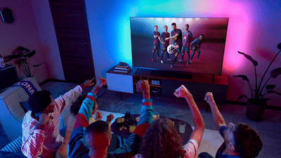 Des amis regardent un match de football sur un téléviseur Philips Ambilight et célèbrent une action de jeu, tandis qu’un éclairage coloré immersif illumine le salon.