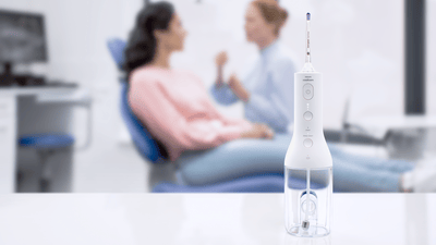 A Philips water flosser is shown clearly stood on a counter in the foreground while a dental professional and patient are shown blurred in the background.