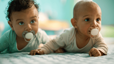 Two babies lay on their stomachs on a soft surface, wearing Philips Avent soothers, and looking away from the camera.