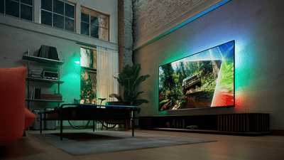 Philips Ambilight TV in a contemporary loft living room, displaying an off-road racing scene with colorful ambient light extending beyond the screen onto the surrounding wall.