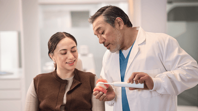 A man wearing a dentist’s scrubs shows a female patient how to brush teeth properly using some dentures and an electric toothbrush.