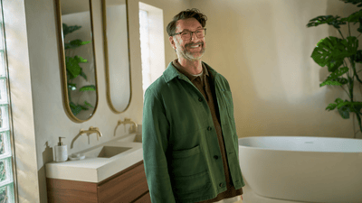 A man with a beard wearing a green jacket and dark glasses is standing in a stylish bathroom and is smiling at the camera.