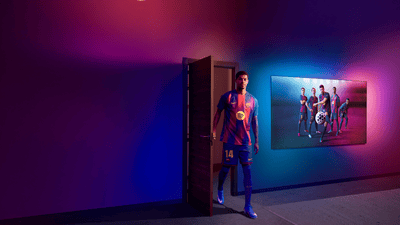 FCB player walks through an open door into a neon-lit room, with a Philips Ambilight TV displaying FCB players on screen.