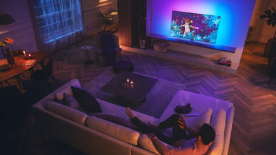 Woonkamerscène met een Philips Ambilight-tv die kleurrijk licht verspreidt terwijl een persoon ontspant op een bank
