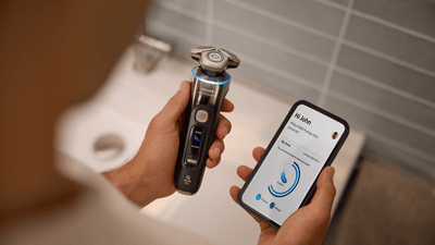 A man looking at the GroomTribe app on his phone while holding the Philips Shaver i9000 Prestige Ultra in the other hand.