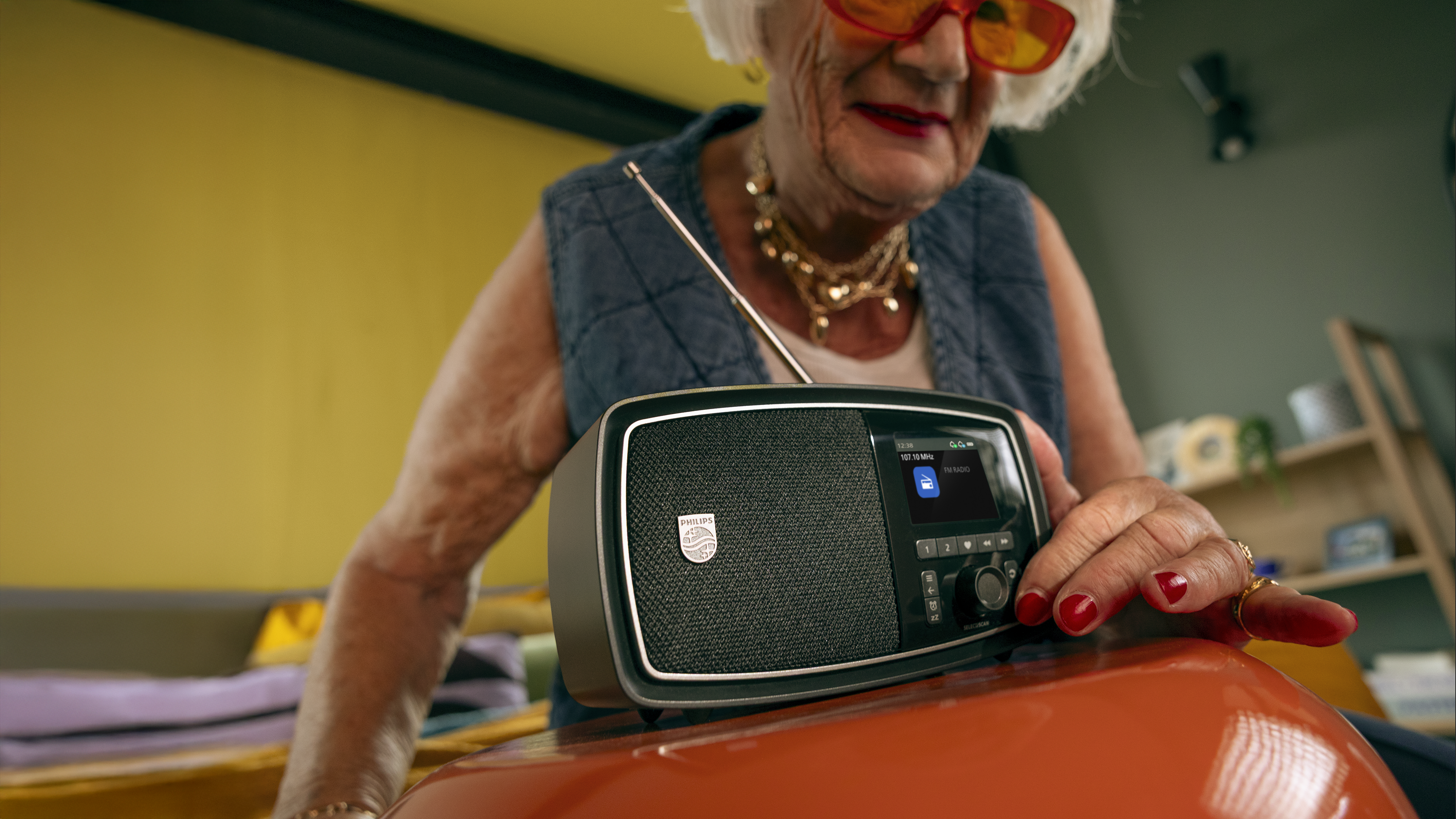 Starejša elegantna ženska v udobnem domačem okolju nežno prilagaja kompakten radio Philips na mizi in uživa v prijetnih trenutkih poslušanja.