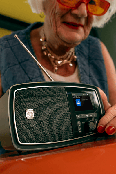 Starejša elegantna ženska v udobnem domačem okolju nežno prilagaja kompakten radio Philips na mizi in uživa v prijetnih trenutkih poslušanja.