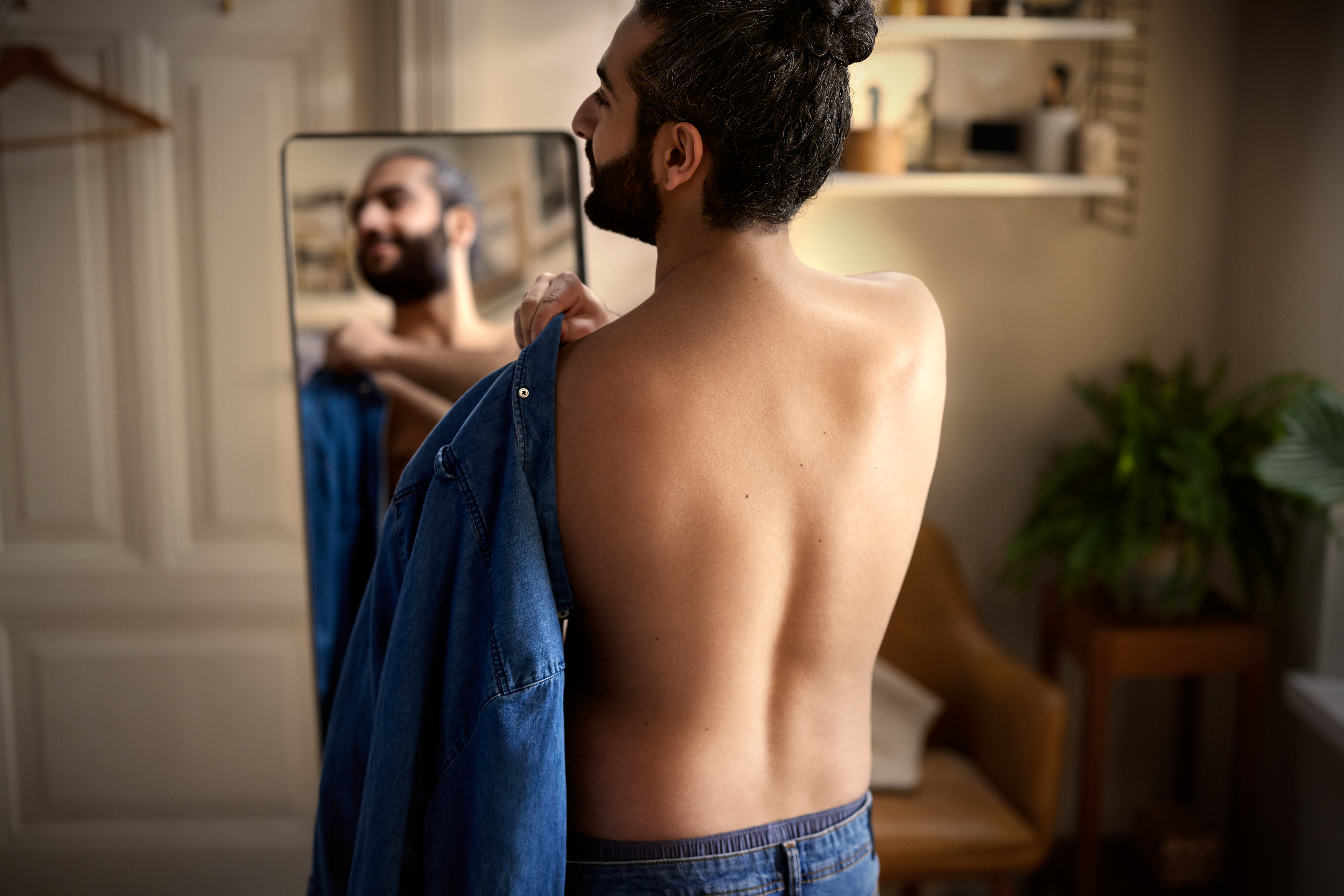 A man puts on a shirt in front of a mirror, his back facing towards the camera.