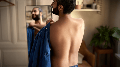 A man puts on a shirt in front of a mirror, his back facing towards the camera.