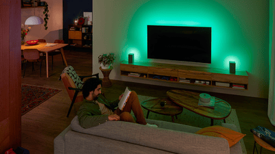 Άτομο που χαλαρώνει σε καναπέ με τηλεόραση Philips Ambilight, πράσινος ατμοσφαιρικός φωτισμός φωτίζει τον τοίχο.