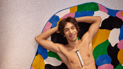 A shirtless young man is lying on a colourful rug with the Philips OneBlade Intimate on his chest.