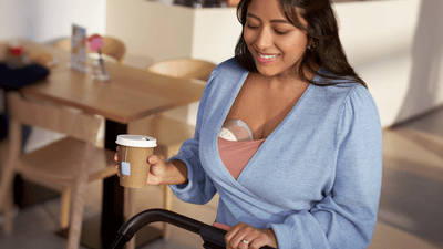 Nuori nainen yllään Philips Avent Natural Care Wearable Breast Pump -rintapumppu pitelee kahvia yhdessä kädessä ja työntää vauvanrattaita toisella.