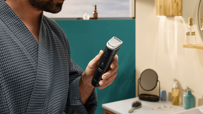 A bearded man in a blue robe holds a black Philips trimmer in a bathroom.