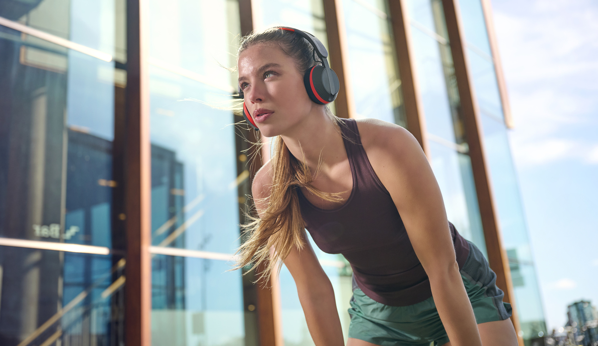 Una donna che indossa le cuffie Philips A6219 si prende una pausa durante un allenamento all'aperto, in piedi di fronte a un moderno edificio in vetro