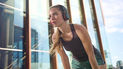 Una donna che indossa le cuffie Philips A6219 si prende una pausa durante un allenamento all'aperto, in piedi di fronte a un moderno edificio in vetro