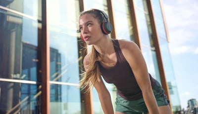 Vrouw met Philips A6219-koptelefoon op neemt een pauze tijdens een buitentraining, staand voor een modern glazen gebouw