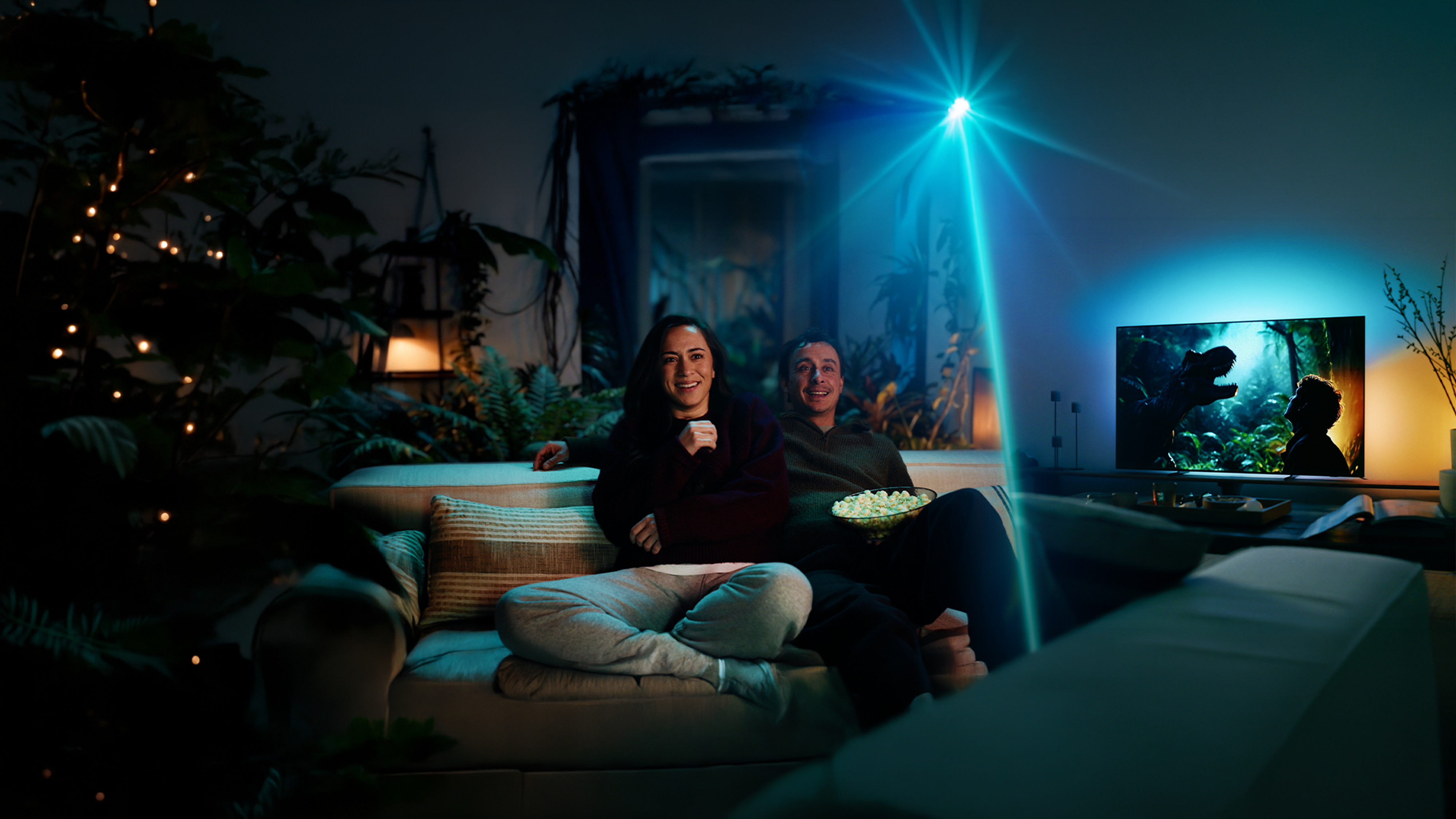 Zwei Personen sitzen auf einem Sofa und sehen auf einen Philips Ambilight TV, dessen stimmungsvolle Hintergrundbeleuchtung das Filmerlebnis in den Raum erweitert.