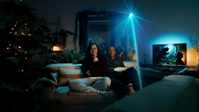 Zwei Personen sitzen auf einem Sofa und sehen auf einen Philips Ambilight TV, dessen stimmungsvolle Hintergrundbeleuchtung das Filmerlebnis in den Raum erweitert.